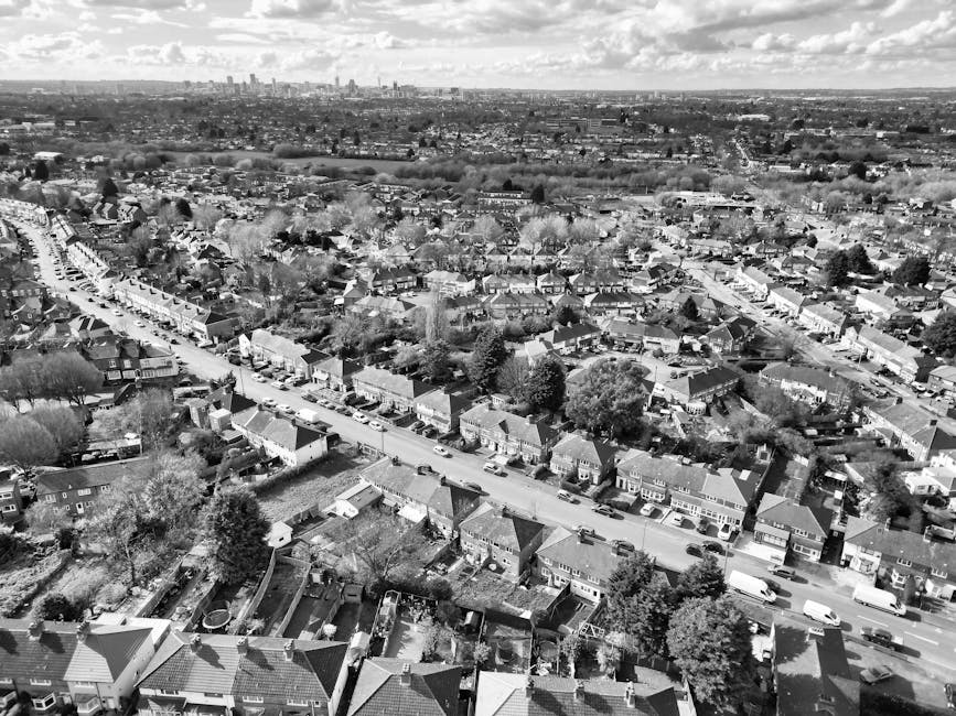 A black and white aerial photograph of a residential neighbourhood showing a row of houses with pitched roofs along a straight street. Cars are parked along the curb on both sides of the road, and some vehicles are in motion. The houses have small front gardens with trees and shrubbery, and some have driveways. In the foreground, there are backyards with fences, sheds, and garden features. The background reveals a distant city skyline with tall buildings, indicating an urban area beyond the suburban houses. The image captures an expansive view of the area, highlighting the density of housing and the layout of streets. This scene may relate to house removals or home relocation services, as provided by Man and a Van Highbury, who assist with furniture transport and packing during moving processes.