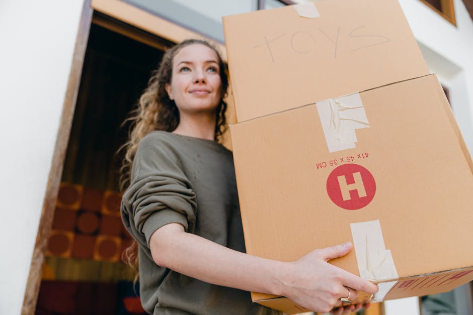 A young woman with curly brown hair and fair skin, wearing a green sweatshirt with rolled-up sleeves, is indoors holding two cardboard boxes during a house relocation. One box has 'TOYS' written on it with a marker, and both are sealed with packing tape. She is standing near a doorway, with a wooden doorframe visible behind her, and a room with colorful patterned wallpaper or decorations in the background. The boxes are positioned in front of her chest, and she is gripping them securely, suggesting careful handling during packing and moving. The natural lighting highlights her composed facial expression, and the environment indicates an organized packing process, typical of professional removals services such as those provided by Man and a Van Highbury, supporting home relocation and furniture transport tasks.