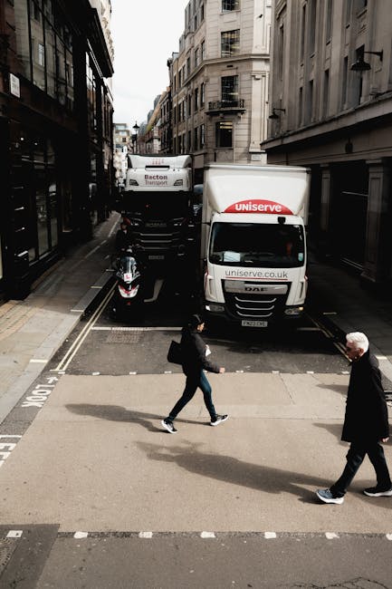 A street scene showing a narrow urban road with tall, multi-story buildings on either side. In the background, a black moving truck and a white delivery van loaded with furniture, boxes, and packing materials are positioned at a loading area near the curb. The moving vans are part of a home relocation process, with the vehicles parked on a designated loading zone. In the foreground, two pedestrians are crossing the road on a pedestrian crossing—one person with dark clothing and a backpack, the other with lighter hair wearing a dark coat. The environment suggests busy city logistics related to house removals, with the vans ready for furniture transport and loading activities. Man and a Van Highbury occasionally handles such logistics, supporting efficient packing and moving services.