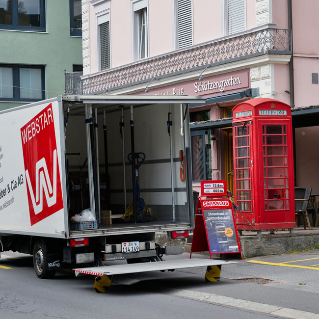 A moving van with an open side panel is parked on a street in Highbury, showing a staged loading area with packing materials, a blue hand truck, and various moving equipment inside. The van is positioned next to a red telephone box on the pavement, with a residential building behind featuring balconies and a sign for a pub and garden. A small table and chairs are visible outside one of the buildings. The scene captures a home relocation or furniture transport process, with the van prepared for loading or unloading of household items, as part of a professional removals service. The setting is well-lit with natural daylight, emphasizing the urban environment and the equipment used in packing and moving households, aligning with local house removals in Highbury offered by Man and a Van Highbury.
