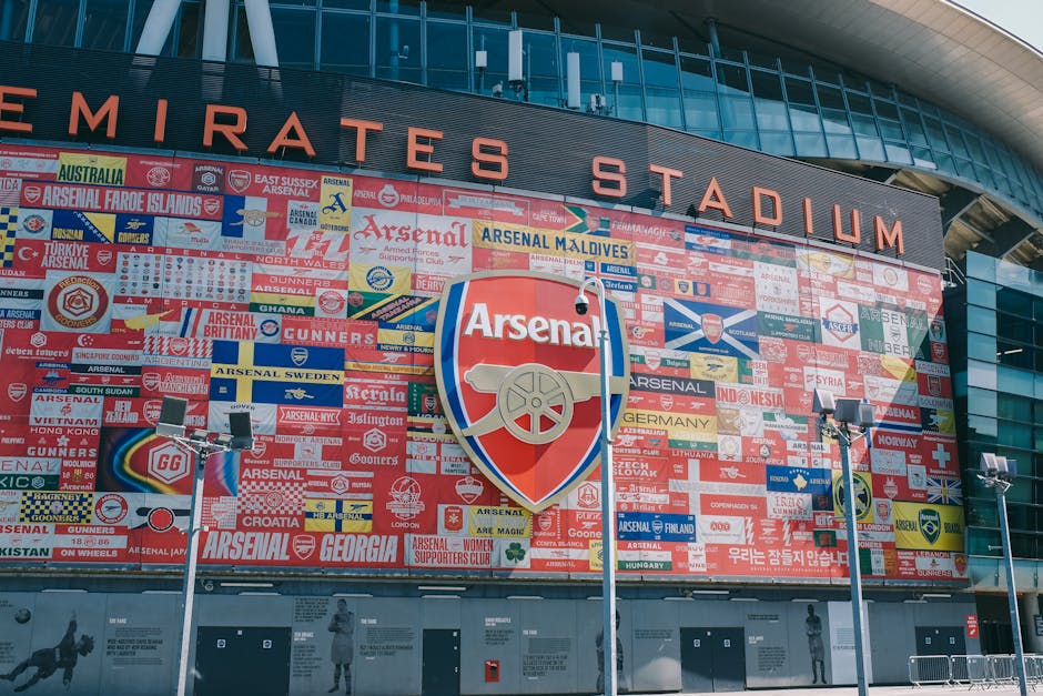 Exterior view of Emirates Stadium with a large mural displaying various flags and symbols associated with Arsenal football club, featuring the prominent Arsenal crest in the center. In front of the stadium, there are three tall portable floodlights positioned on the pavement, which is free of vehicles or traffic. The stadium's façade is made of glass and metal, with large windows and a curved roof extending overhead. The scene is captured during daylight with bright, natural lighting highlighting the architectural features of the stadium. This image appears to relate to house removals or moving services by providing context on large-scale transport and logistics, where the stadium serves as an example of a significant building that may require careful furniture transport and loading processes by experts like Man and a Van Highbury, especially considering any relocation or temporary equipment setup near large venues.