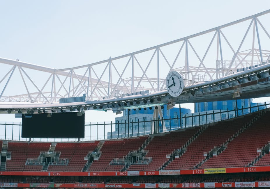 Exterior view of Emirates Stadium with a large mural displaying various flags and symbols associated with Arsenal football club, featuring the prominent Arsenal crest in the center. In front of the stadium, there are three tall portable floodlights positioned on the pavement, which is free of vehicles or traffic. The stadium's façade is made of glass and metal, with large windows and a curved roof extending overhead. The scene is captured during daylight with bright, natural lighting highlighting the architectural features of the stadium. This image appears to relate to house removals or moving services by providing context on large-scale transport and logistics, where the stadium serves as an example of a significant building that may require careful furniture transport and loading processes by experts like Man and a Van Highbury, especially considering any relocation or temporary equipment setup near large venues.