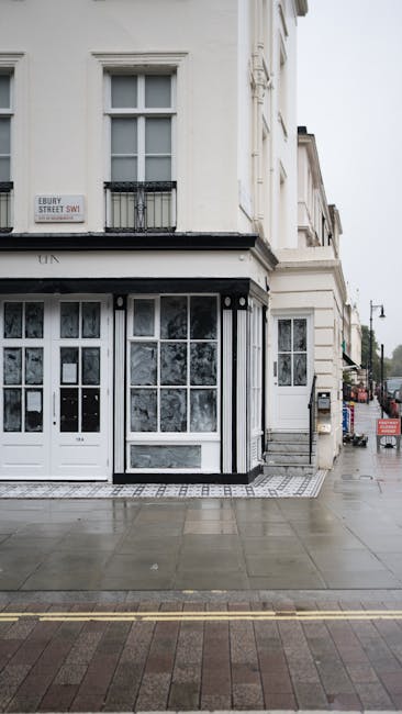 A photograph of the exterior corner of a three-storey white building on Ebury Street in Highbury, London, showing large ground-floor windows with white frames and decorative glass panels, and a black iron balcony on the first floor. The pavement in front is wet, likely from recent rain, with visible reflections on the surface. To the right, a black van is parked on the street while a stacking trolley loaded with cardboard boxes and packing materials is positioned near the building entrance, indicating a home relocation or furniture transport process. The image captures part of the adjacent street with other buildings and trees in the distance, under an overcast sky. The scene reflects an urban environment suitable for house removals and packing services provided by Man and a Van Highbury.