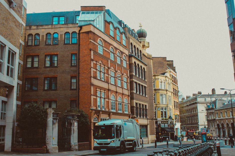 A street view of a multi-storey brick building with large arched windows, surrounded by other historic and modern buildings in an urban area. A white van from Man and a Van Highbury is parked on the pavement near a gated entrance to the building, which has stone pillars and greenery. The van is positioned close to the entrance, suggesting a home or office relocation process, with the vehicle and nearby bollards indicating active loading or unloading activities. The background shows additional buildings with diverse architectural styles, including a pale yellow structure with decorative facades, and a clear sky overhead. This setting reflects a typical scene where furniture and packing materials may be loaded or unloaded during moving services as part of a comprehensive house or office removal in Highbury.