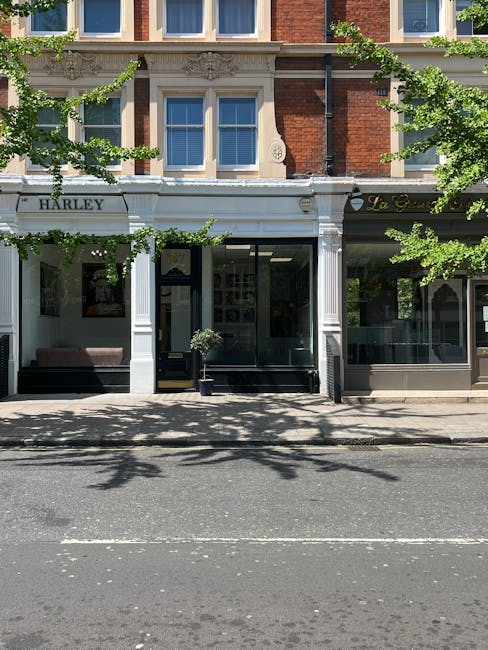 A photograph of the exterior corner of a three-storey white building on Ebury Street in Highbury, London, showing large ground-floor windows with white frames and decorative glass panels, and a black iron balcony on the first floor. The pavement in front is wet, likely from recent rain, with visible reflections on the surface. To the right, a black van is parked on the street while a stacking trolley loaded with cardboard boxes and packing materials is positioned near the building entrance, indicating a home relocation or furniture transport process. The image captures part of the adjacent street with other buildings and trees in the distance, under an overcast sky. The scene reflects an urban environment suitable for house removals and packing services provided by Man and a Van Highbury.