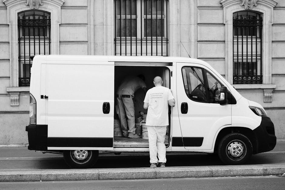 A person wearing a blue long-sleeved shirt and a black padded vest is seen standing beside an open moving van with a partially visible rear door. Inside the van, several cardboard boxes of varied size