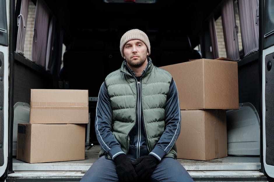 A young man with a serious expression, wearing a gray knitted beanie, a green puffer vest, navy blue track jacket, and black gloves, sitting inside the open back of a cargo van. The van’s interior contains several cardboard boxes of various sizes, some stacked on top of each other, wrapped in plastic or packing materials. The boxes are positioned on the vehicle’s metal flooring, with part of the van’s dark interior visible behind him. The scene appears to be part of a home relocation or furniture transport process, with the man possibly preparing for or completing a moving task as part of a professional removals service. The lighting is natural, indicating daytime, and the setting is at the driveway or loading area adjacent to a residential property, aligned with the services provided by Man and a Van Highbury.