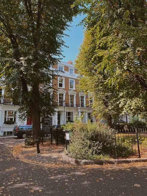 A multi-storey white residential building on Highbury Park seen through large green trees with dense foliage, casting shadows on the pavement below. In front of the building, there is a black wrought 